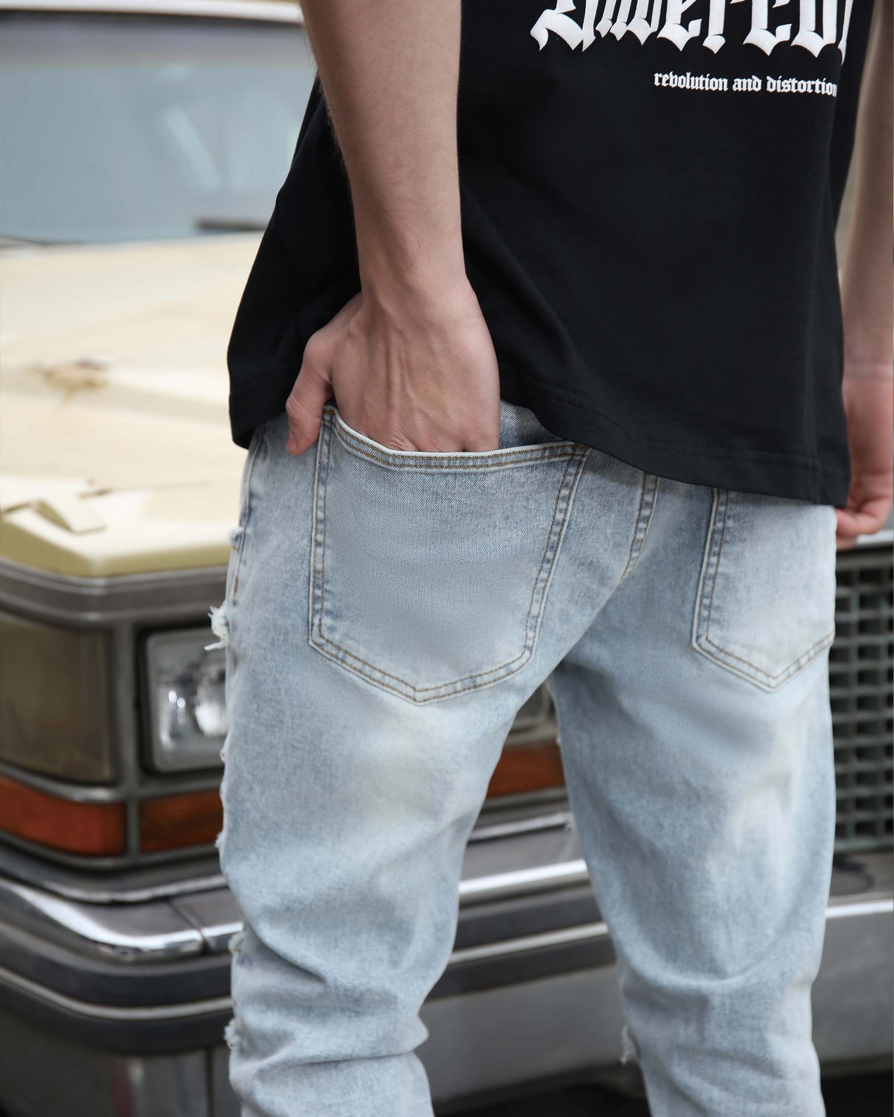 Man wearing LOGEQI black tee & light blue stacked patch jeans, hand in back pocket, vintage car, turned back, close-up.