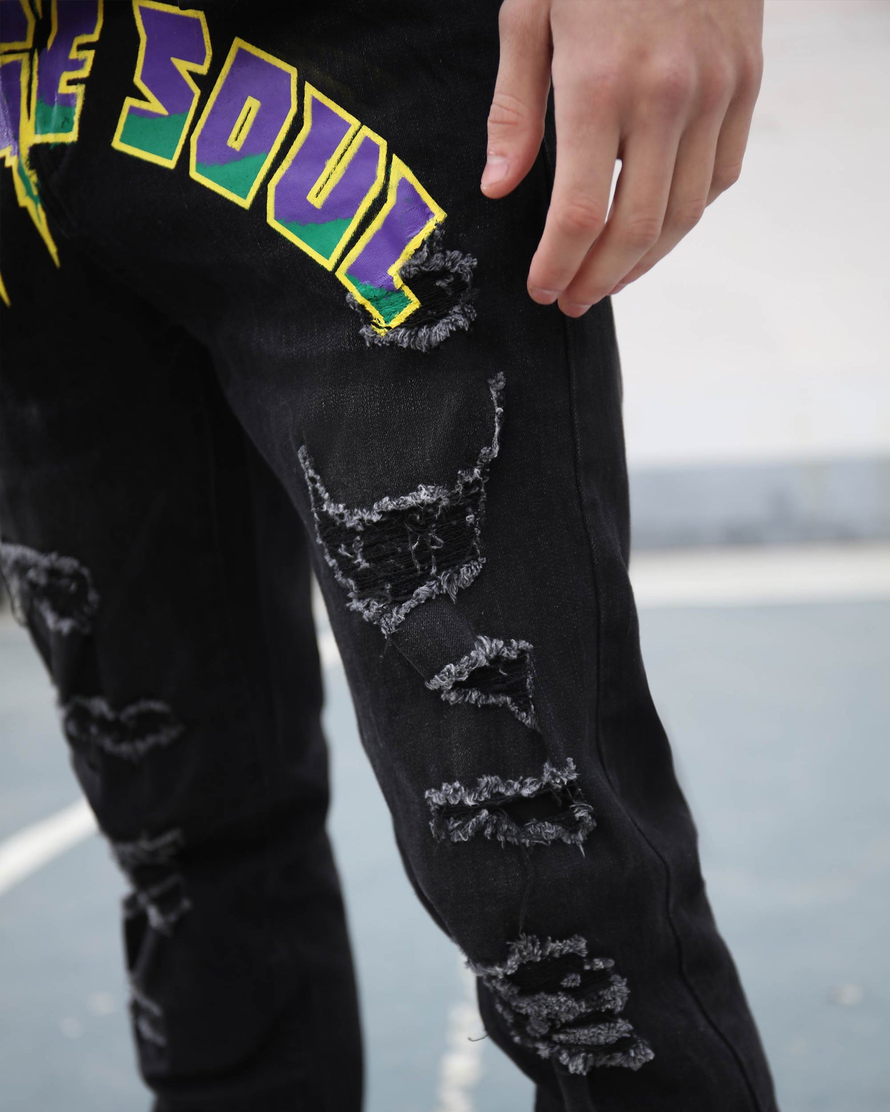 Close-up of LOGEQI black ripped and distressed jeans with colorful “SOUL” print and a hand touching them.