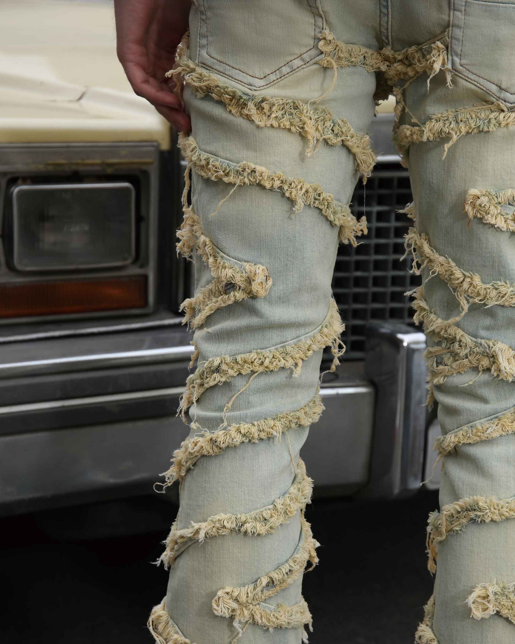 Close-up of LOGEQI's men's light-wash vintage distressed jeans with frayed, twisted fabric strips. A hand rests on the hip, with a vintage car in the background