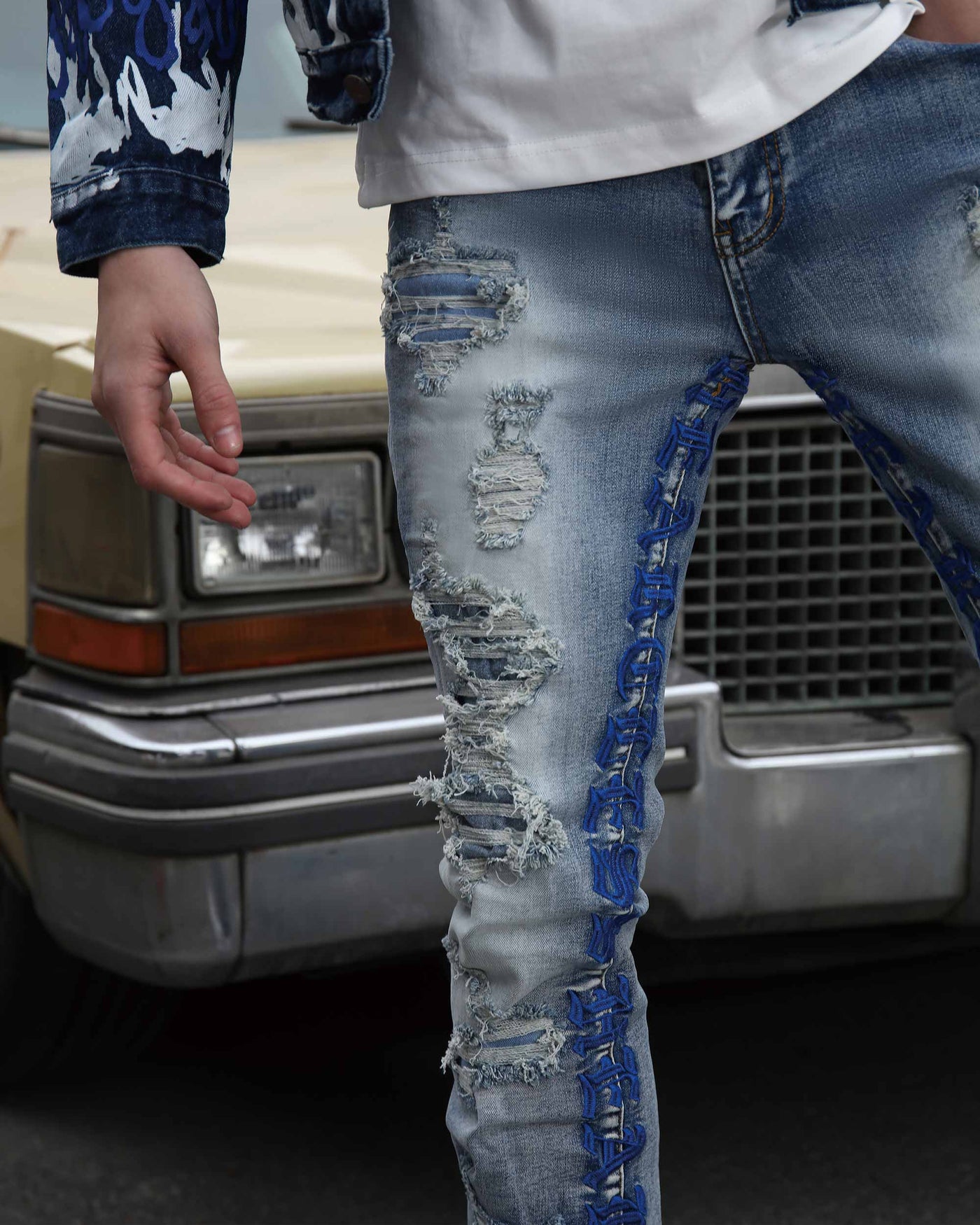 Close-up of light blue ripped jeans with blue embrioderey and patches, paired with a white shirt, near a classic car.