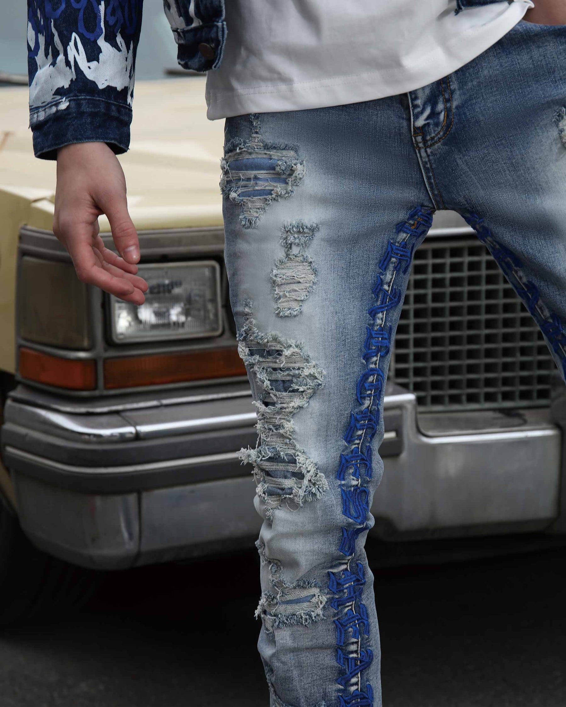 Close-up of light blue ripped jeans with blue embrioderey and patches, paired with a white shirt, near a classic car.