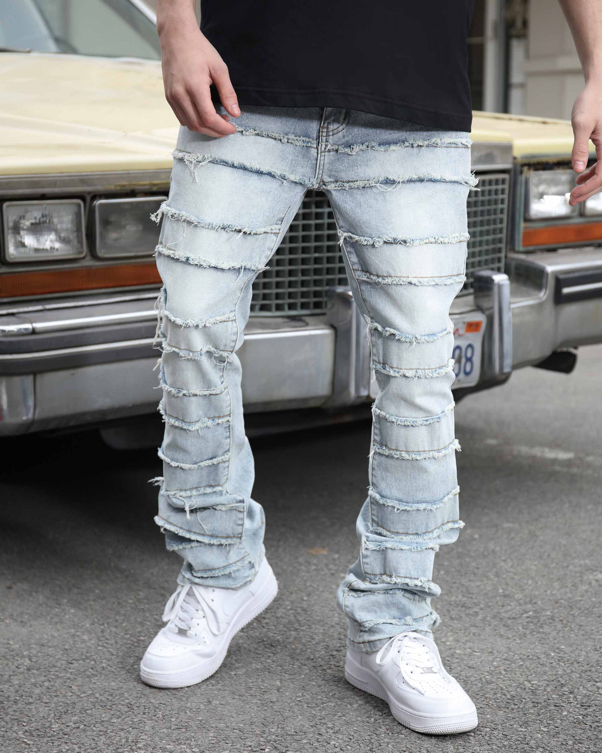 A man wearing a black shirt, light blue stacked-ripped denim jeans with frayed horizontal layers, and white sneakers, standing in front of a vintage car, showcasing streetwear fashion