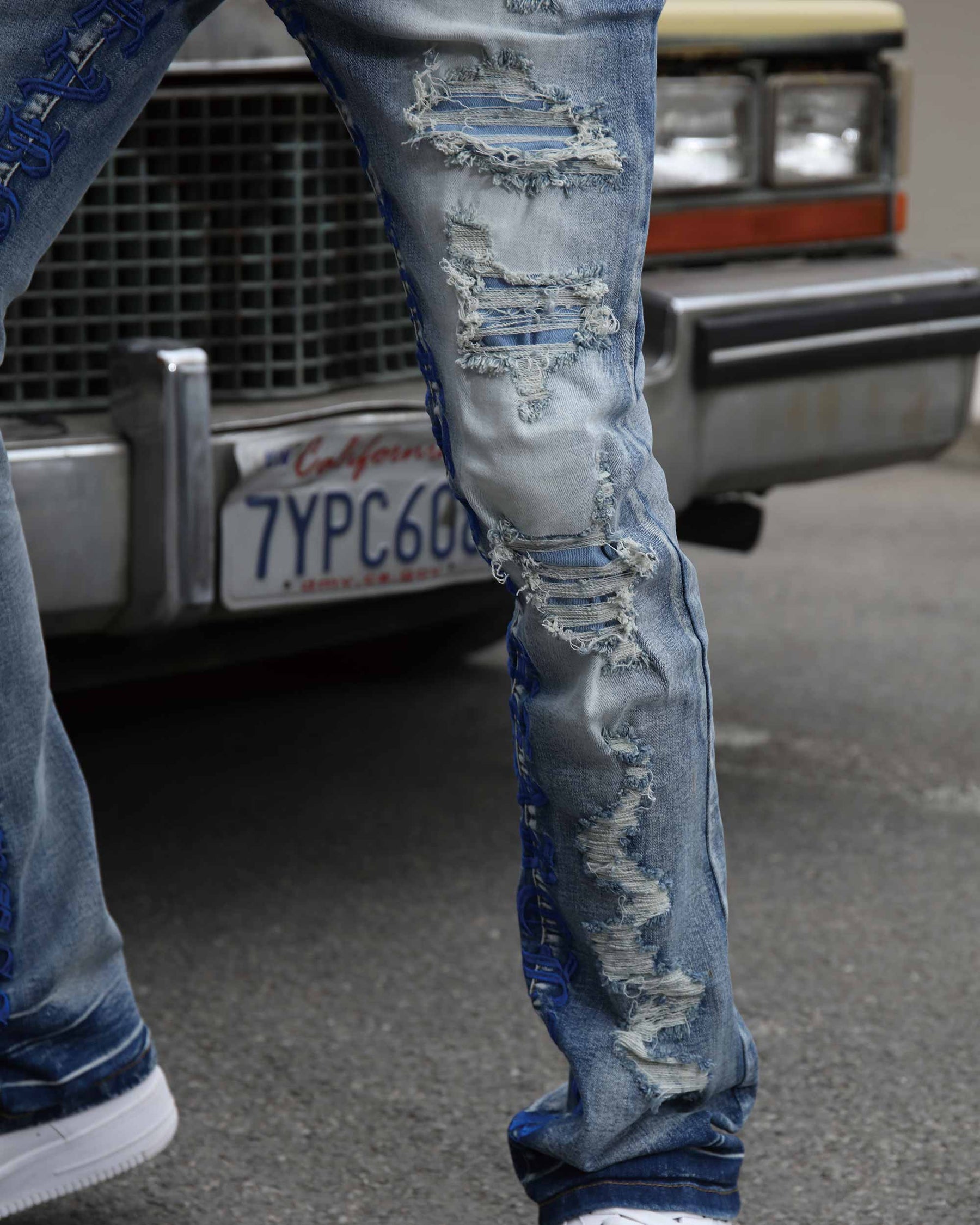 Close-up of LOGEQI men's light blue distressed and ripped patch jeans with blue embroidery, paired with white sneakers, by a vintage car