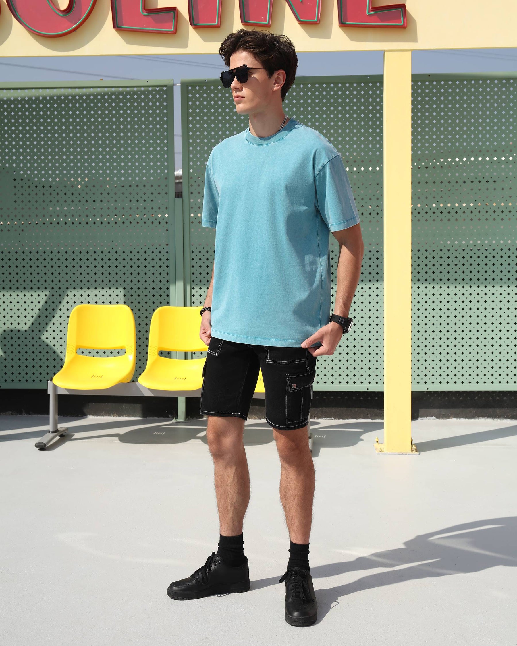 Pairing LOGEQI's denim cargo shorts in black color with a blue t-shirt, sunglasses, and black shoes while walking on the street.