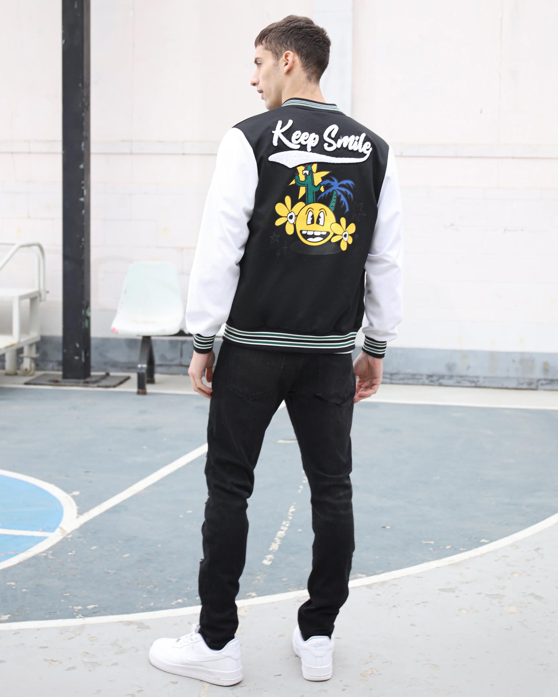 A man in black varsity jacket with “Keep Smile” and graphic, LOGEQI black jeans, white sneakers on a court.