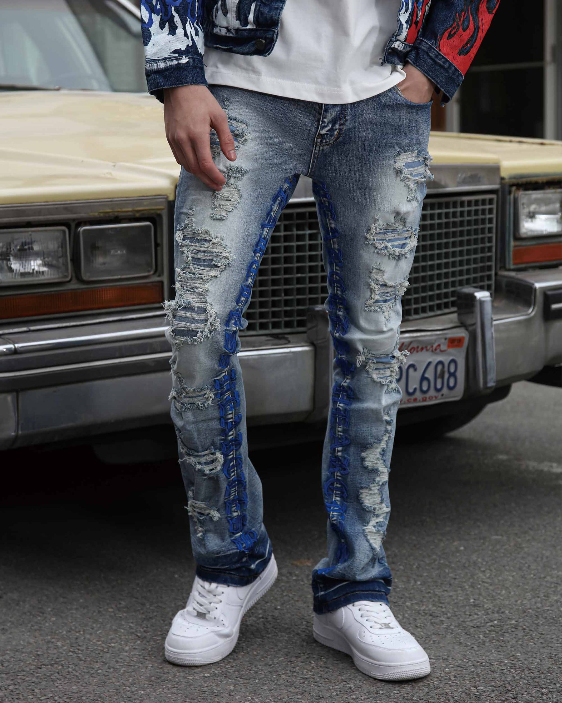 A man wearing LOGEQI's distressed blue patched jeans with blue embroidery leters, a white shirt, a denim jacket, and white sneakers by a vintage car