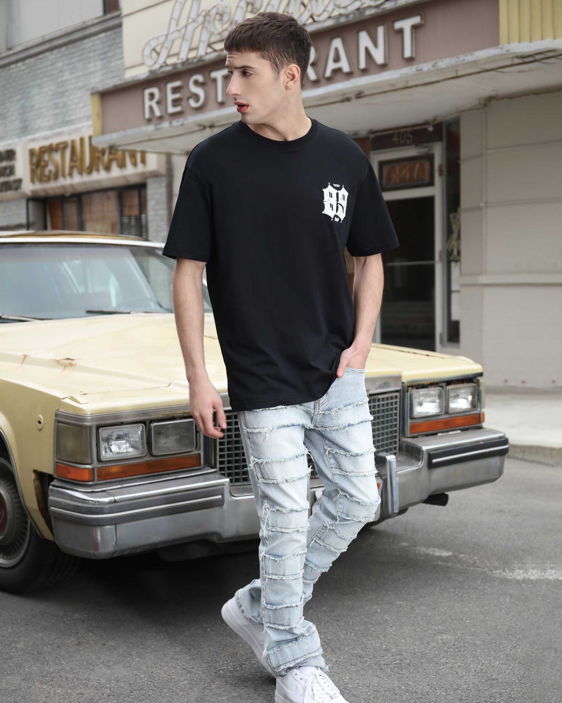 A man in a black tee, LOGEQI's black distressed jeans with layered design and stacked patch design, and white sneakers stands by a vintage car outside a restaurant.