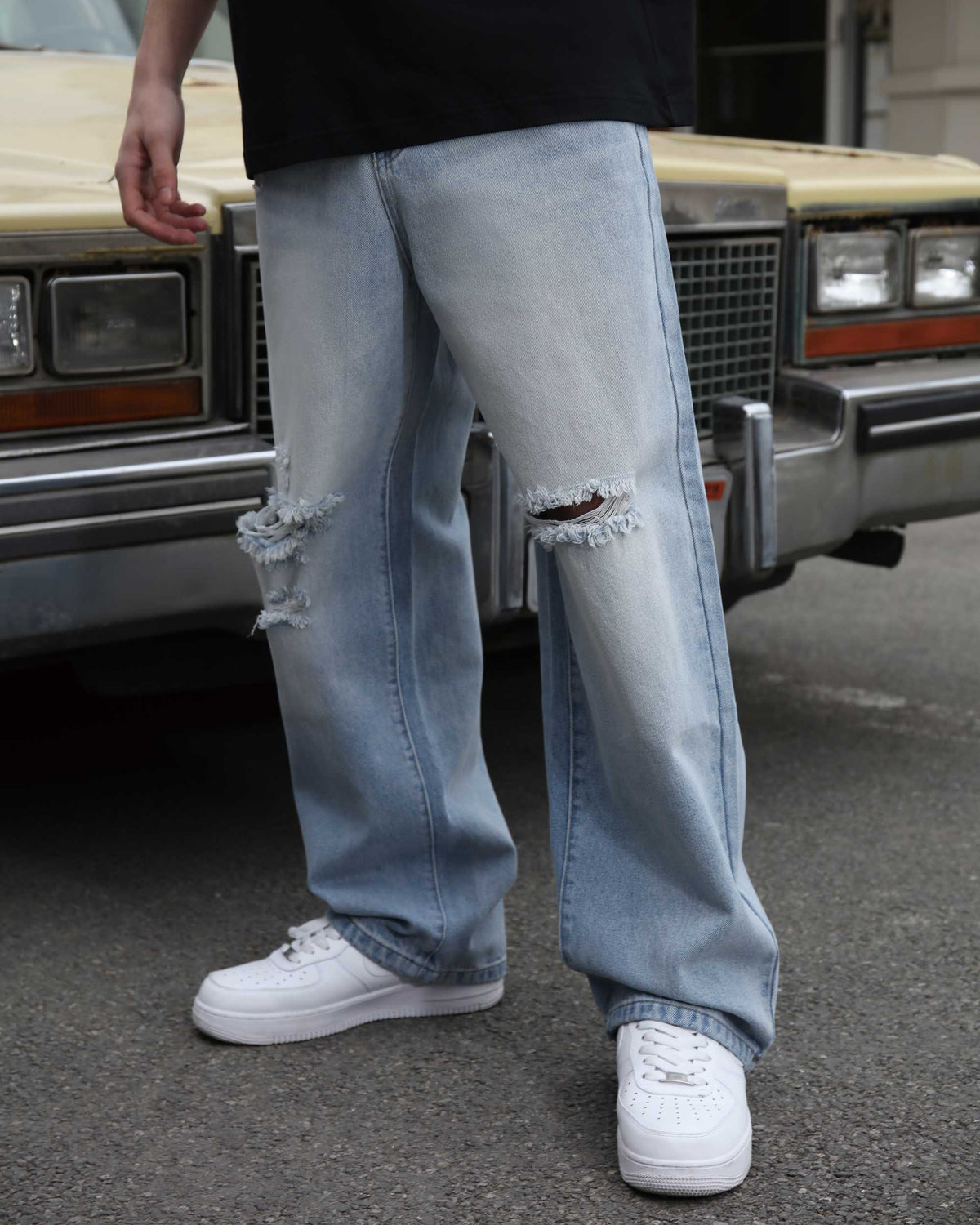 A man showing the side view of LOGEQI's blue ripped baggy jeans with knee rips, standing in front of a car