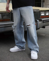 A man showing the side view of LOGEQI's blue ripped baggy jeans with knee rips, standing in front of a car