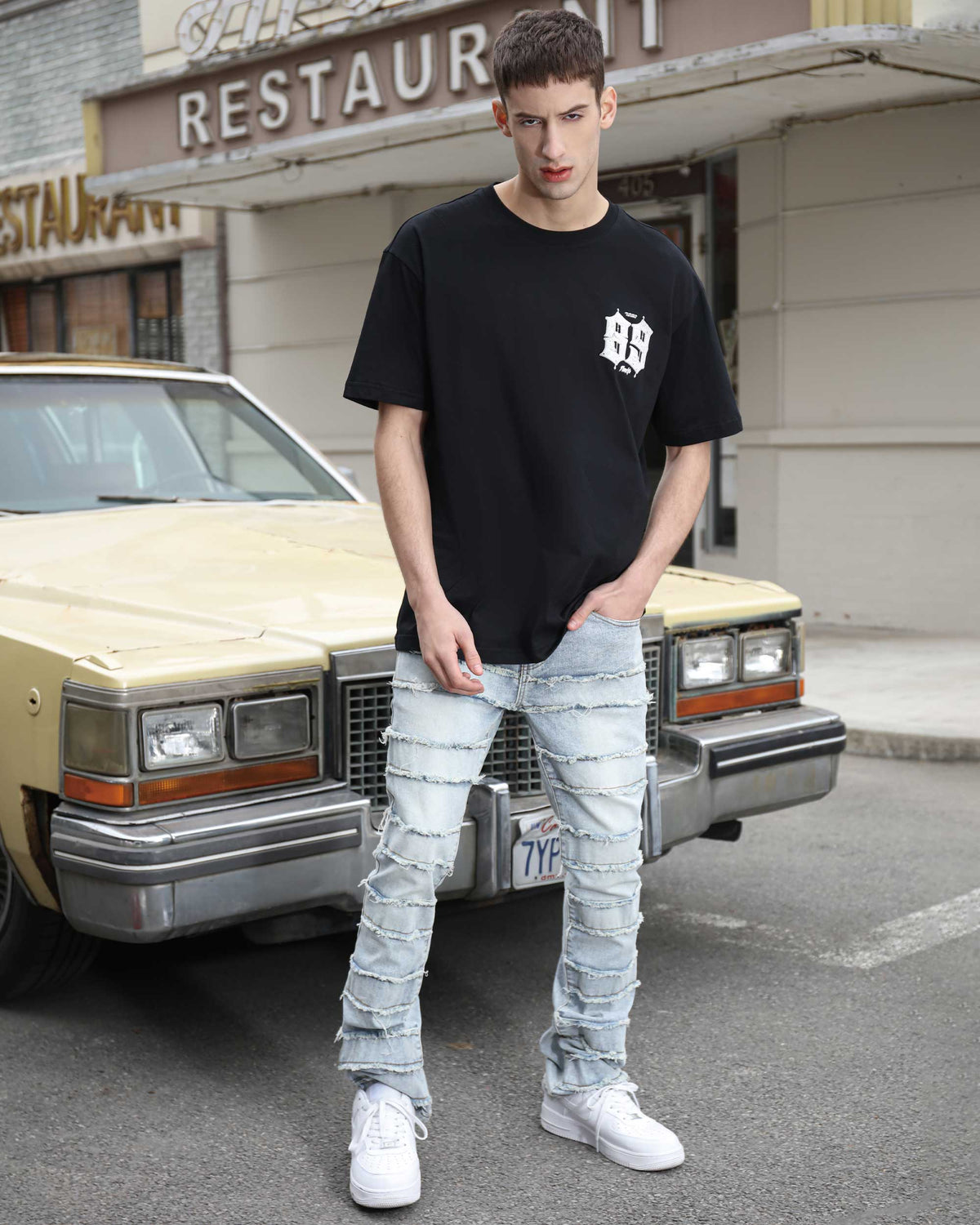 A man stands by a vintage car, wearing a LOGEQI black logo tee, light blue stacked-ripped jeans, and white sneakers, showcasing urban streetwear style
