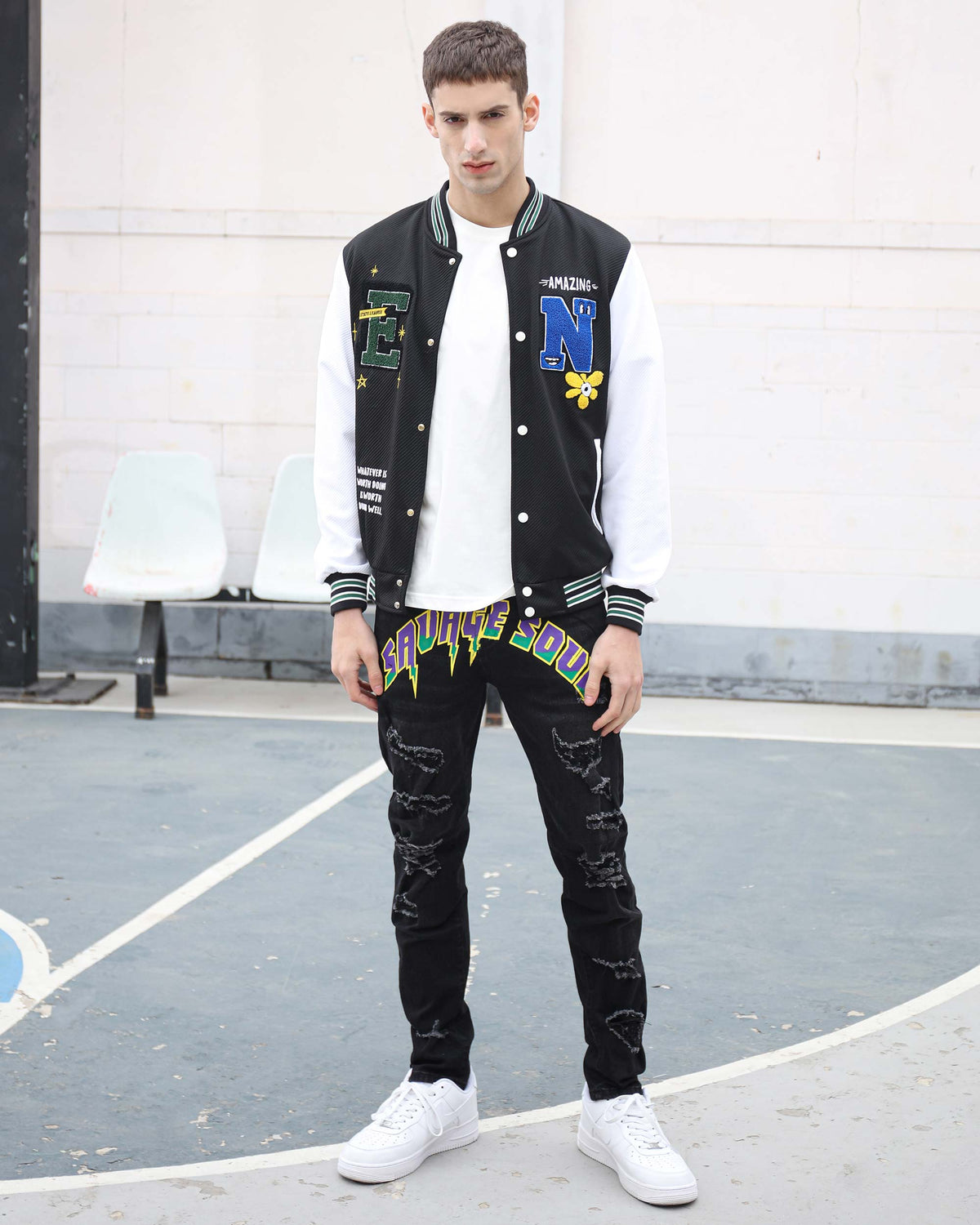 Man standing on an outdoor basketball court wearing LOGEQI black slightly ripped slim jeans with “SAVAGE SOUL” graphic, paired with a black and white varsity jacket with green accents, and white sneakers