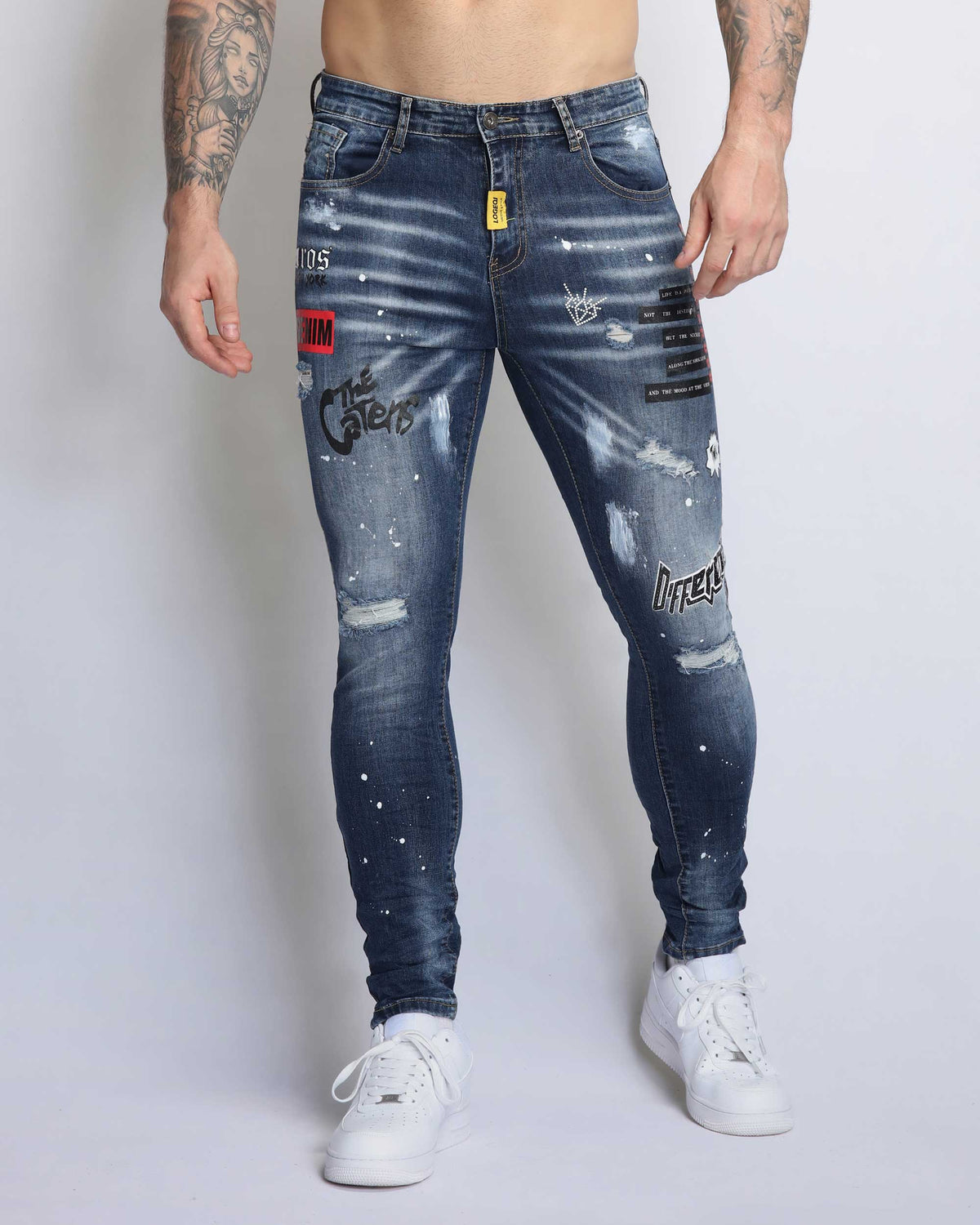 Full-body view of a shirtless man wearing LOGEQI's blue, distressed, and ripped jeans with graffiti-style patches and delicate splatters, paired with white sneakers, showcasing a streetwear style.