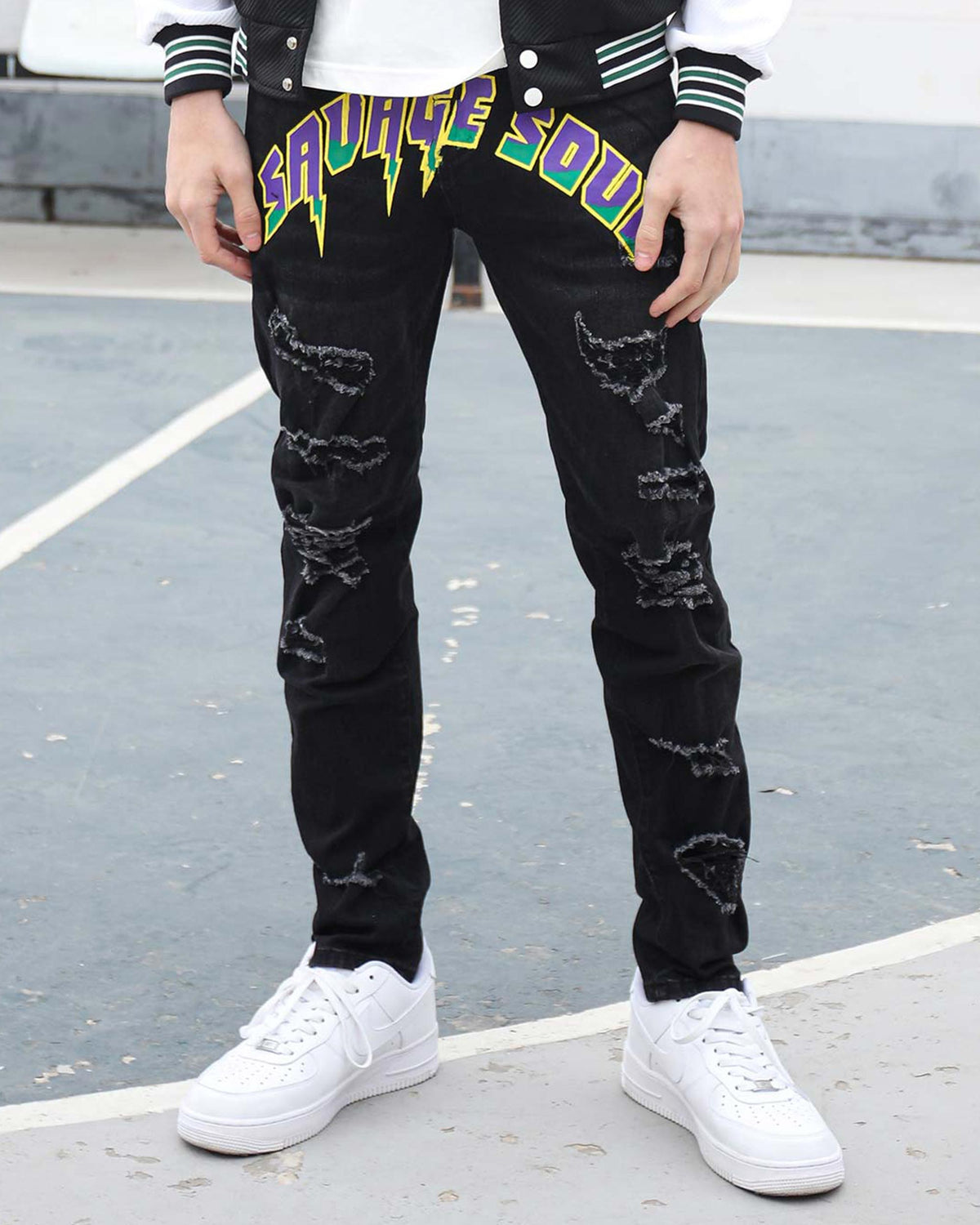 A man wearing LOGEQI black ripped slim-fit jeans with bold “SAVAGE SOUL” graffiti in purple, green, and yellow, styled with white Nike Air Force 1s and a varsity jacket