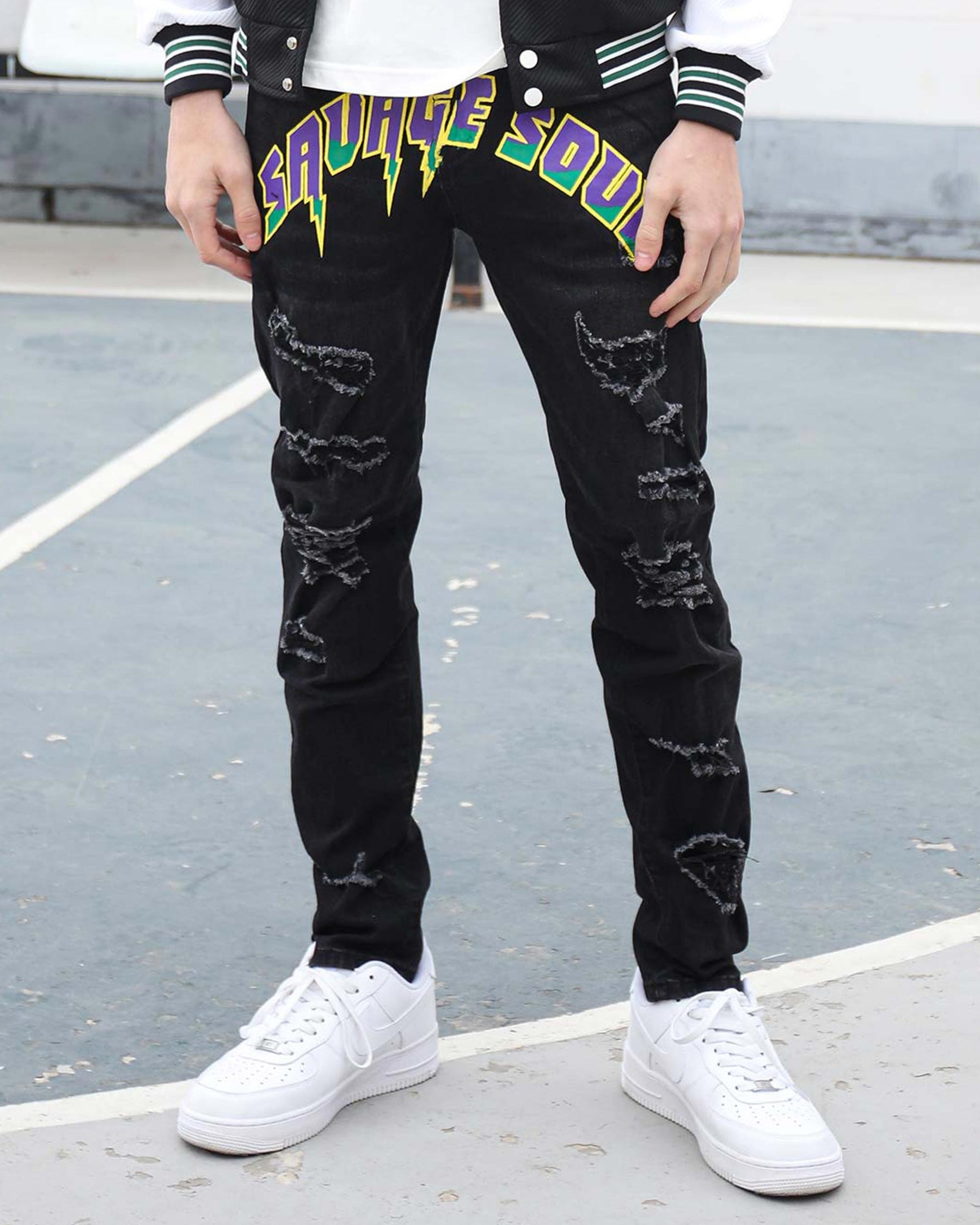 A man wearing LOGEQI black ripped slim-fit jeans with bold “SAVAGE SOUL” graffiti in purple, green, and yellow, styled with white Nike Air Force 1s and a varsity jacket