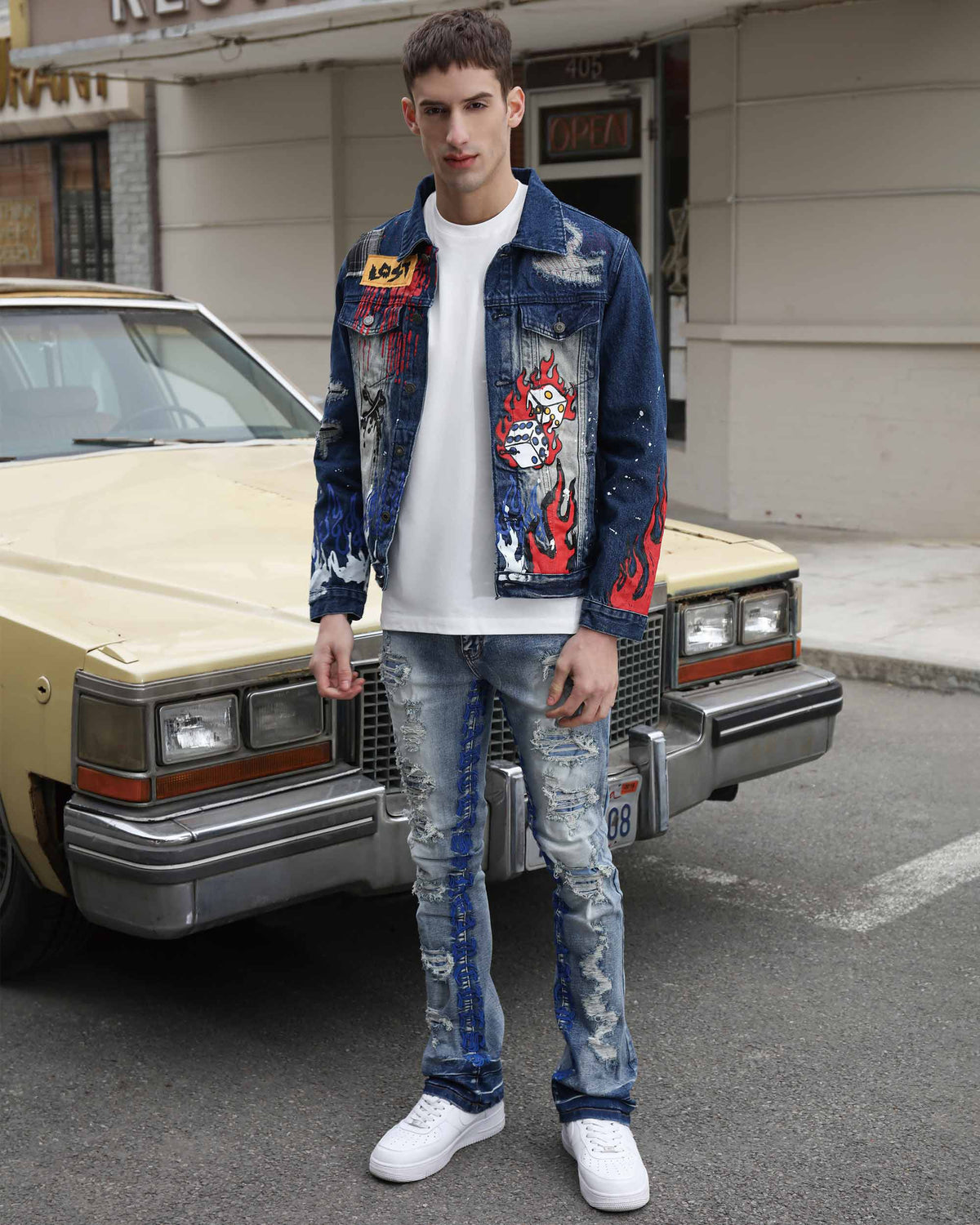 Man in a LOGEQi's distressed blue denim jacket, white tee, blue ripped jeans with patches and embroidered letters, and white sneakers by a vintage car.