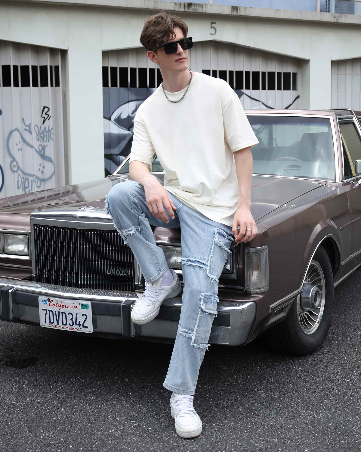 A man wearing LOGEQI's fringed-edge, ripped, and distressed blue baggy jeans is sitting on the hood.