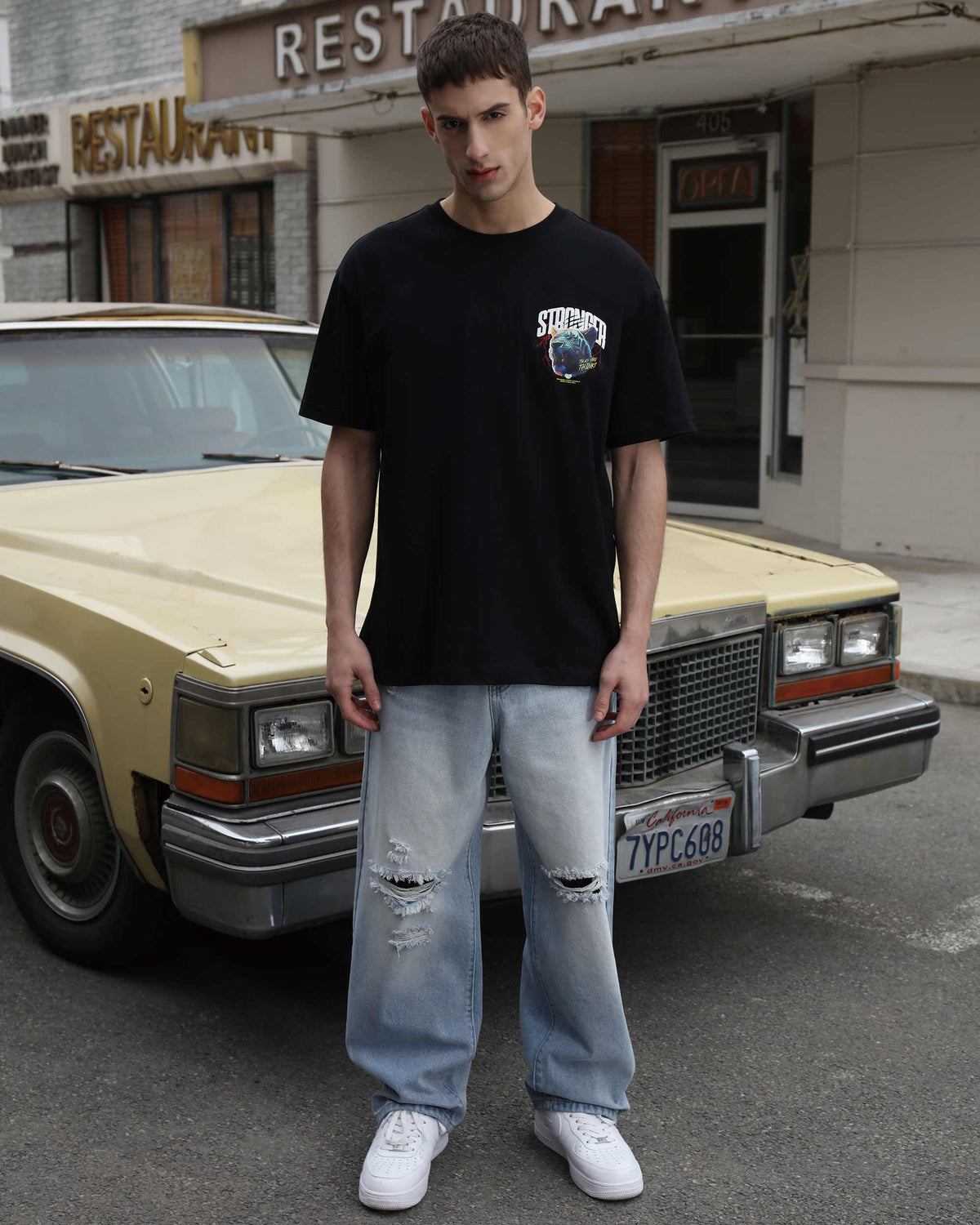 A man wearing blue ripped baggy jeans in a loose fit, paired with a black T-shirt, standing in front of a car on the street.