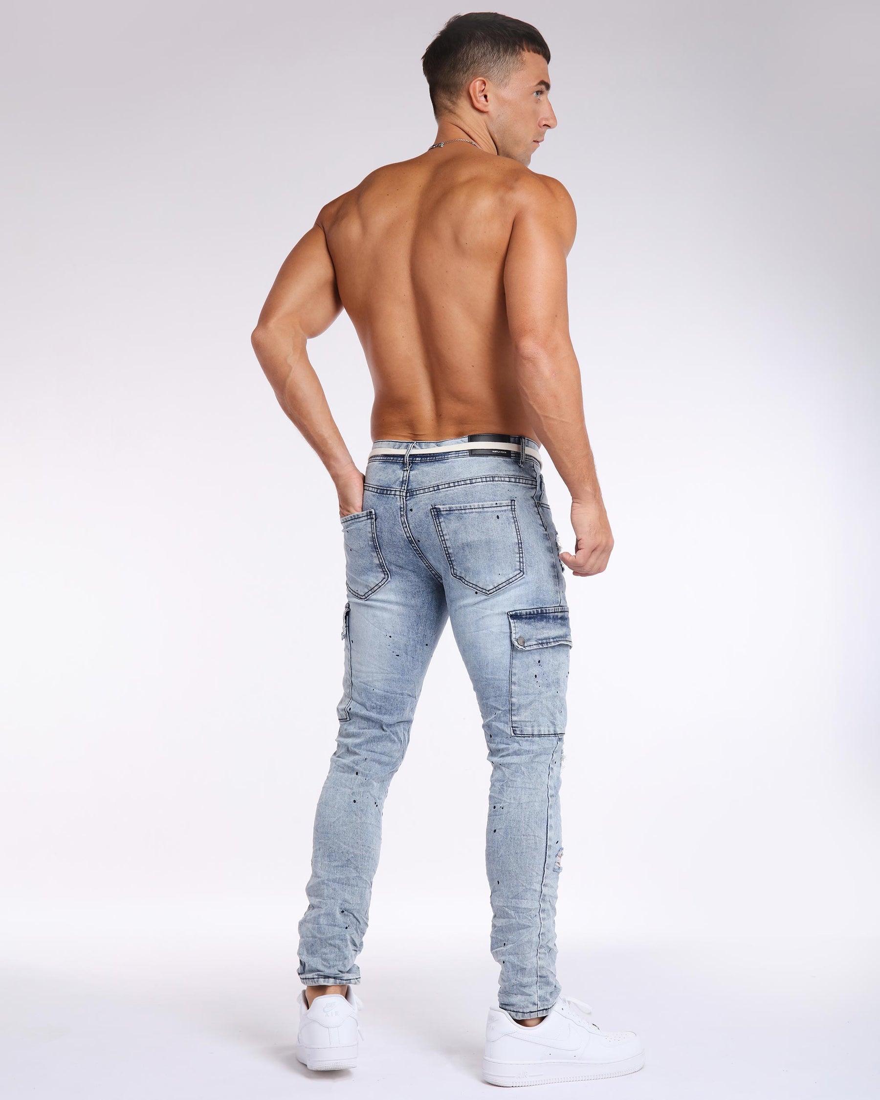 Muscular shirtless man with short black crew-cut, wearing LOGEQI light blue ripped patched jeans with a white strap belt and white shoes, standing with his back facing, head turned, right hand in pocket