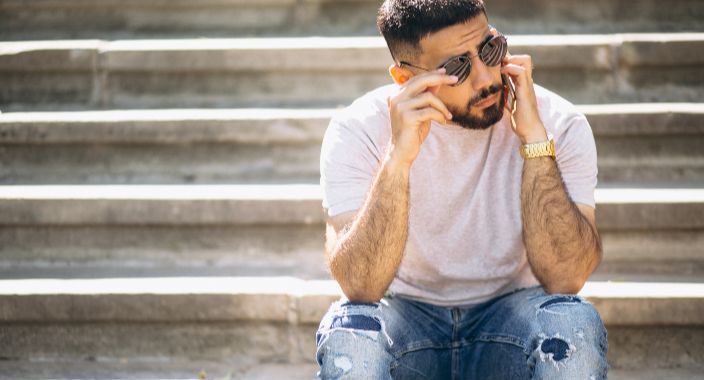 A young, handsome man wearing blue ripped jeans, a white T-shirt, and sunglasses is sitting on the stairs